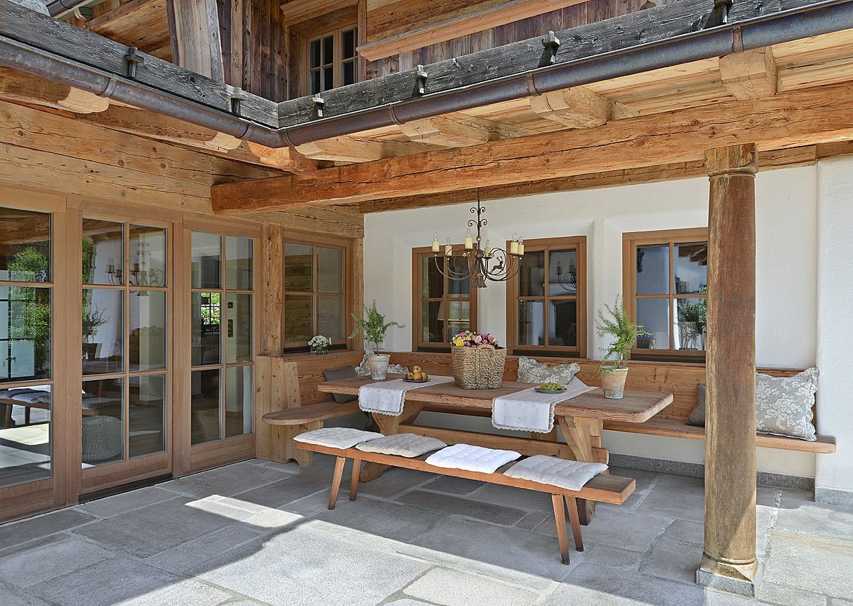 Covered patio with wooden benches, stone flooring, and large windows in a rustic house