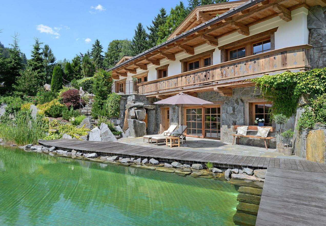 Lakeside chalet with stone patio, wooden balconies, and clear green water surrounded by trees