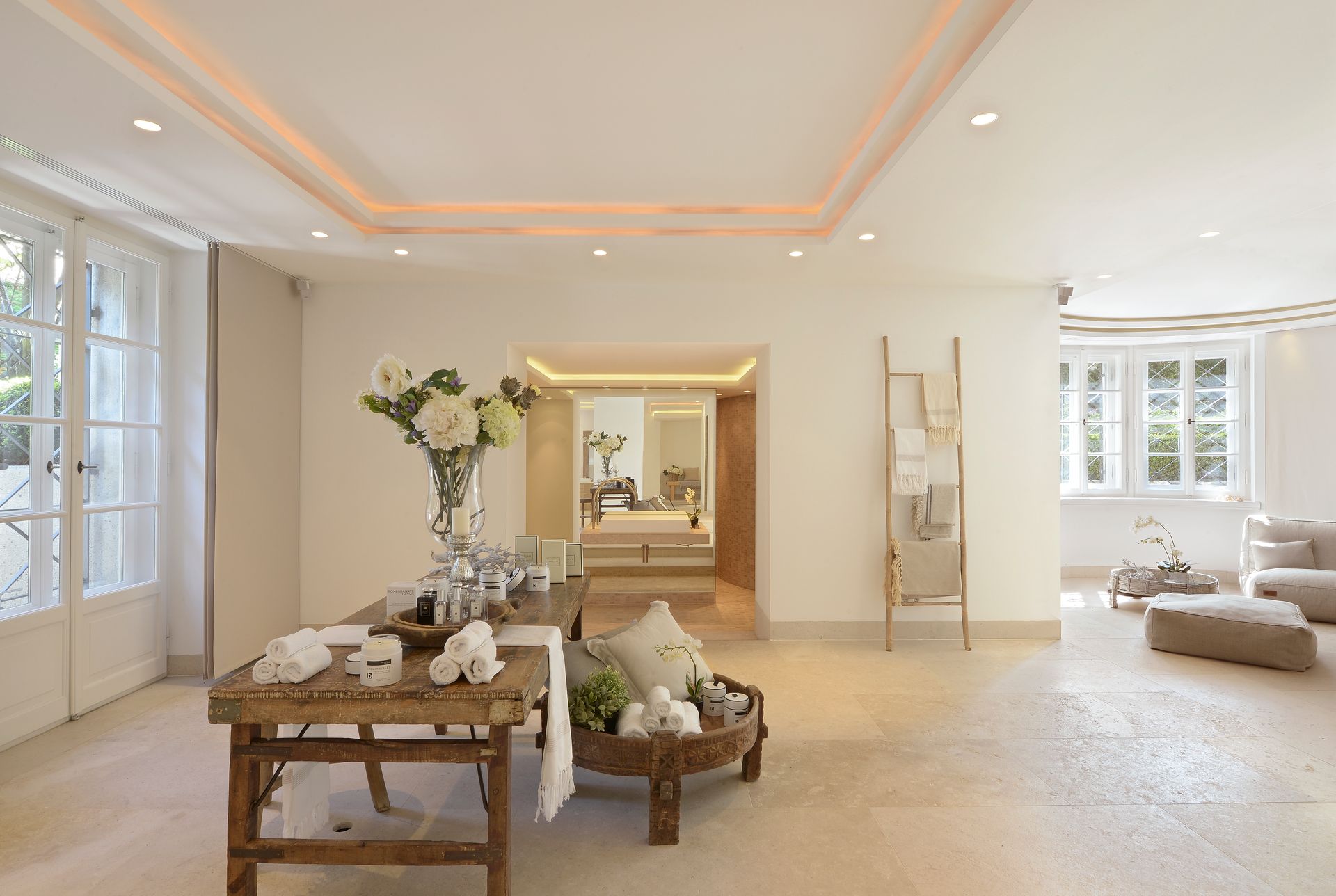 Bright, airy living room with white decor, wooden table, flowers, and large windows