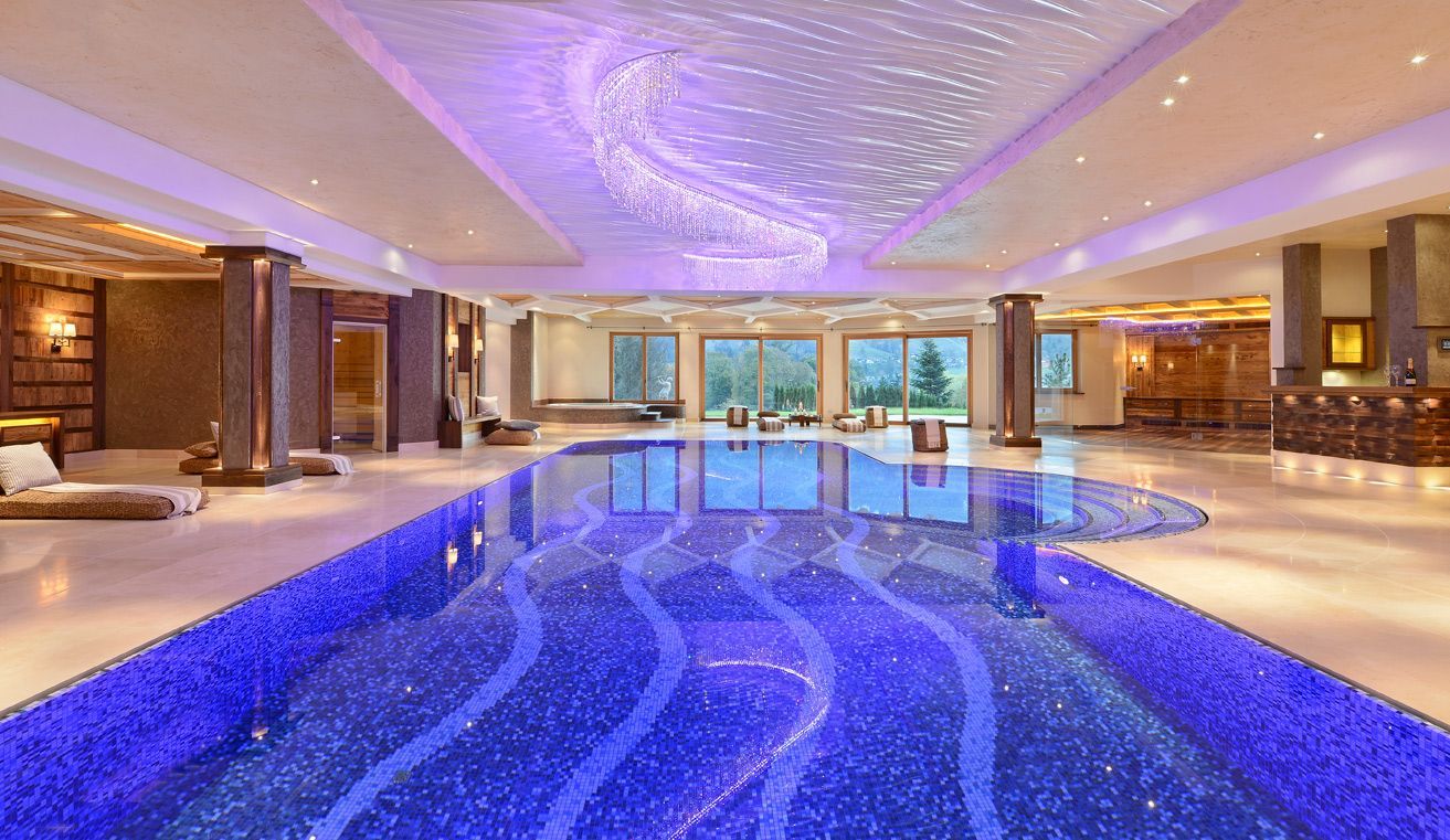 Luxurious indoor pool with blue-lit water, lounge chairs, and glass doors overlooking greenery