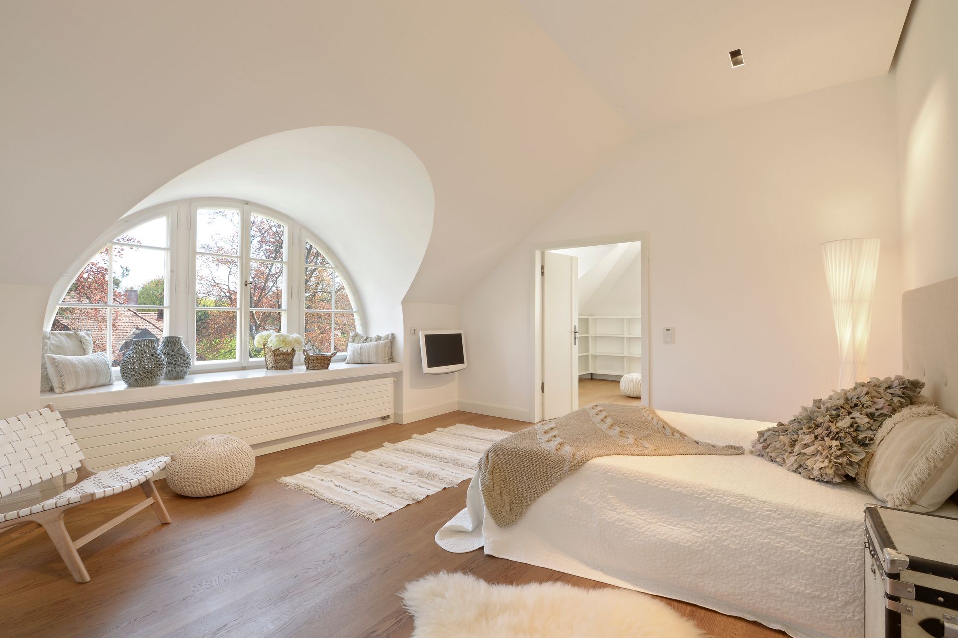 Bright airy bedroom with arched window, white bedding, rug, and wicker chair