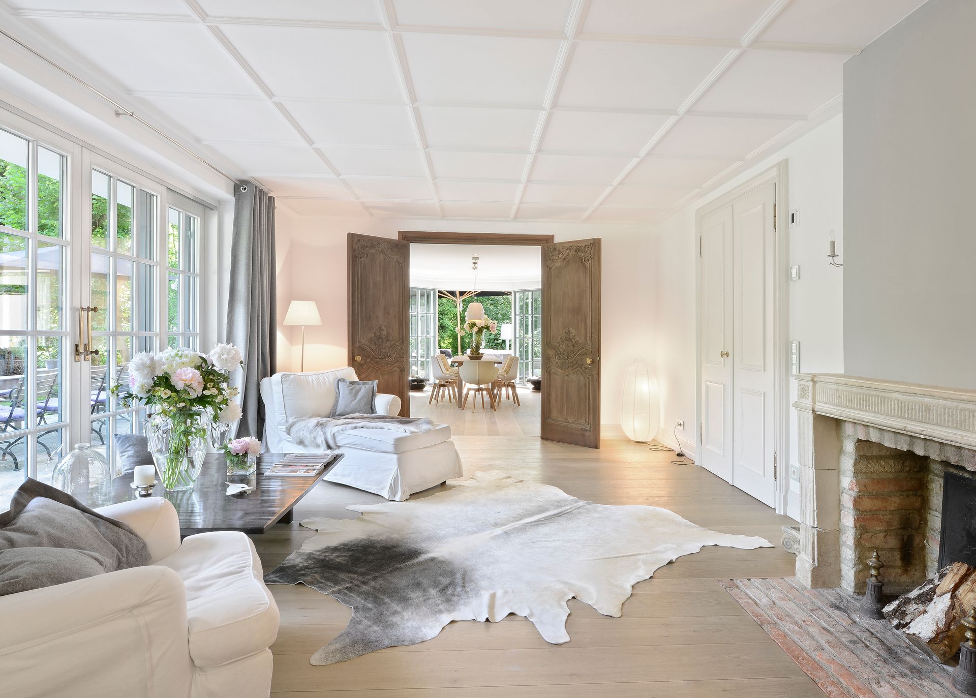 Bright elegant living room with white furniture, large windows, and a fireplace.