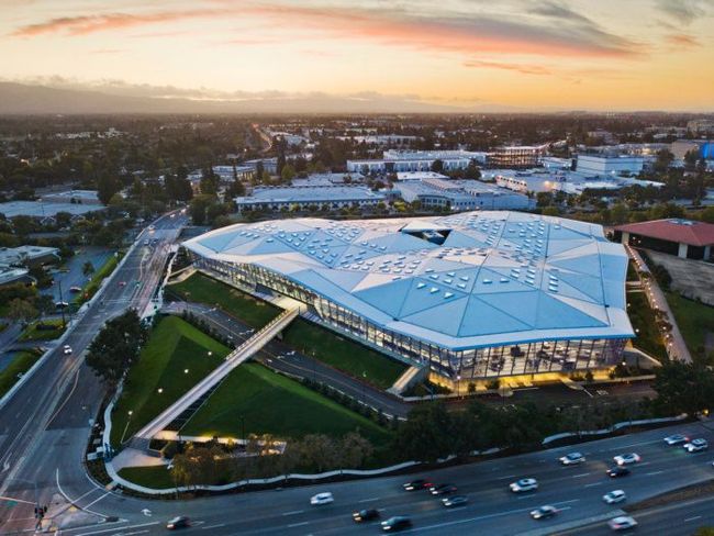 Aerial view of a large modern sports stadium with a white roof at sunset, beside busy roads and parking lots