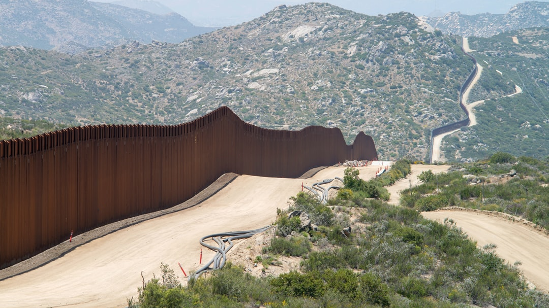 A border fence is surrounded by mountains and a dirt road.