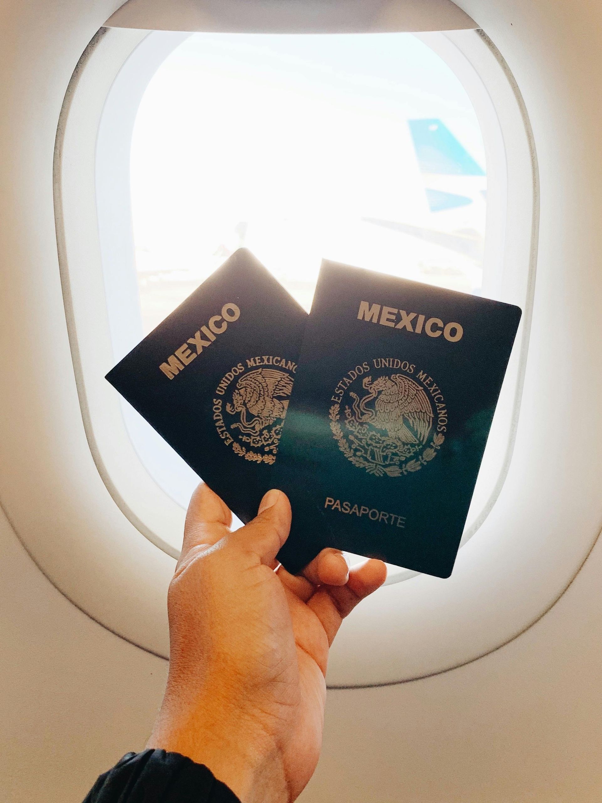 Hand holding two Mexican passports in front of an airplane window.