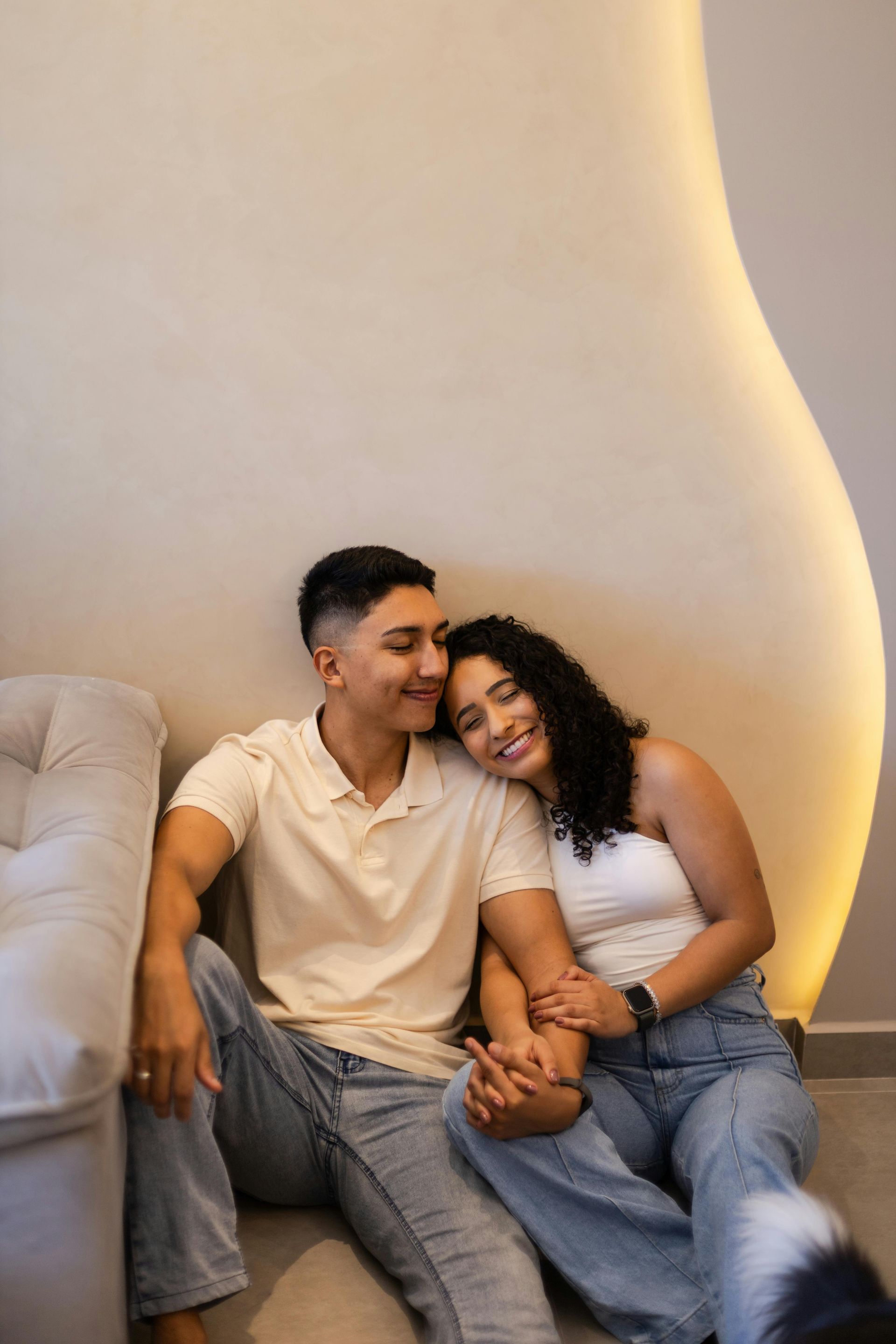 Couple sits together, smiling, holding hands. Woman's head rests on man's shoulder near beige wall.