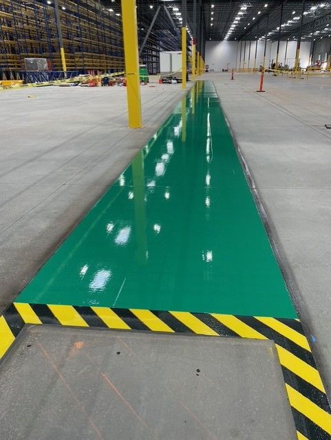Green floor marking in a warehouse aisle, bordered by black-and-yellow hazard stripes, with yellow support columns.