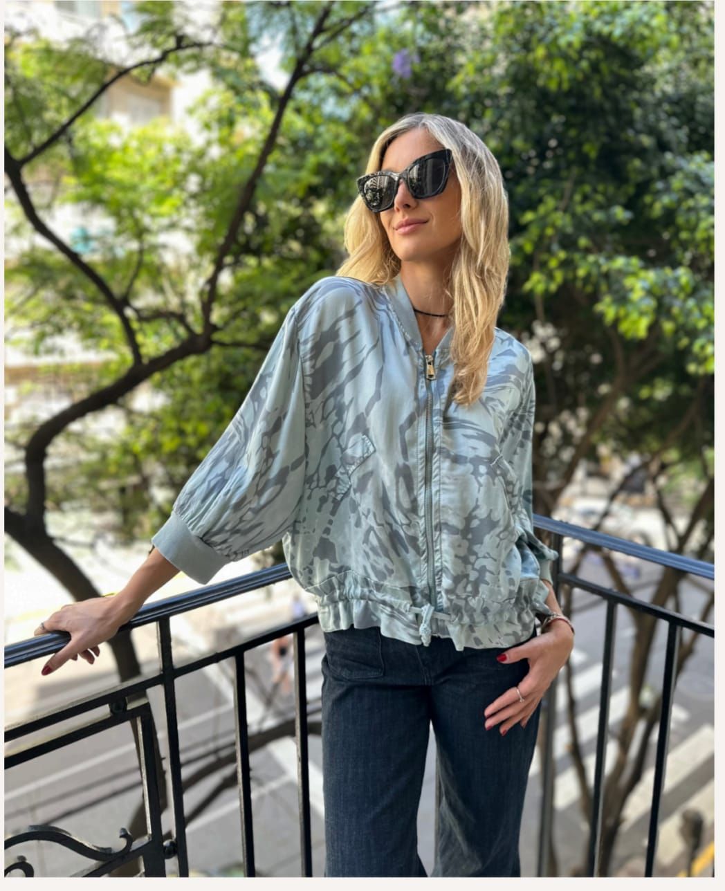 Mujer con gafas de sol en un balcón, vestida con una blusa azul estampada y vaqueros, con árboles al fondo.