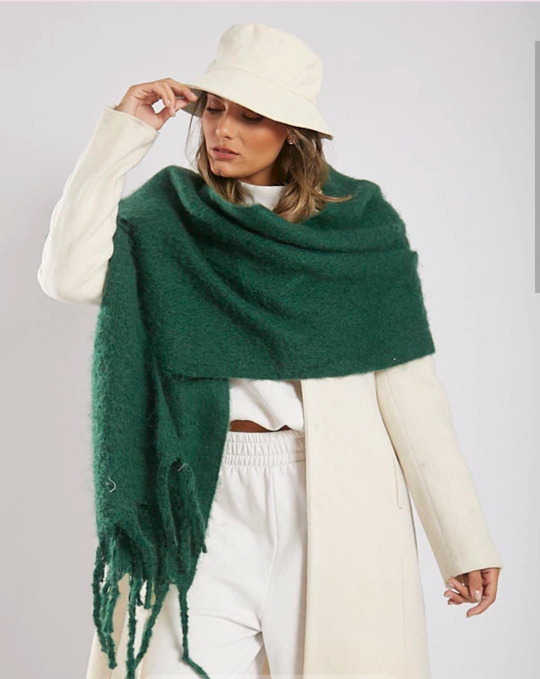 Mujer con sombrero y abrigo color crema y una gran bufanda verde, posando sobre un fondo claro.