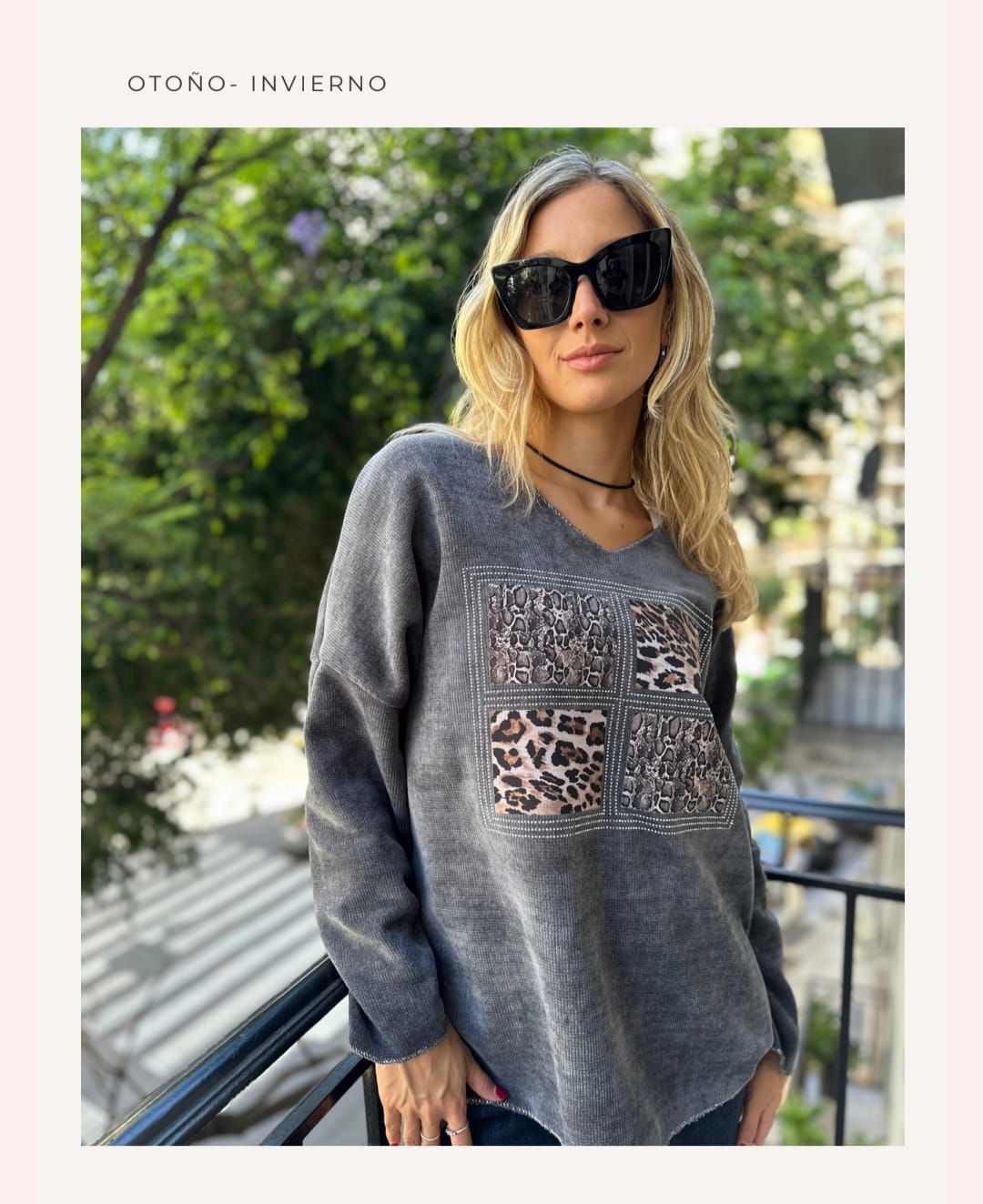 Mujer con gafas de sol y un jersey gris con estampado de leopardo, de pie al aire libre en un balcón.