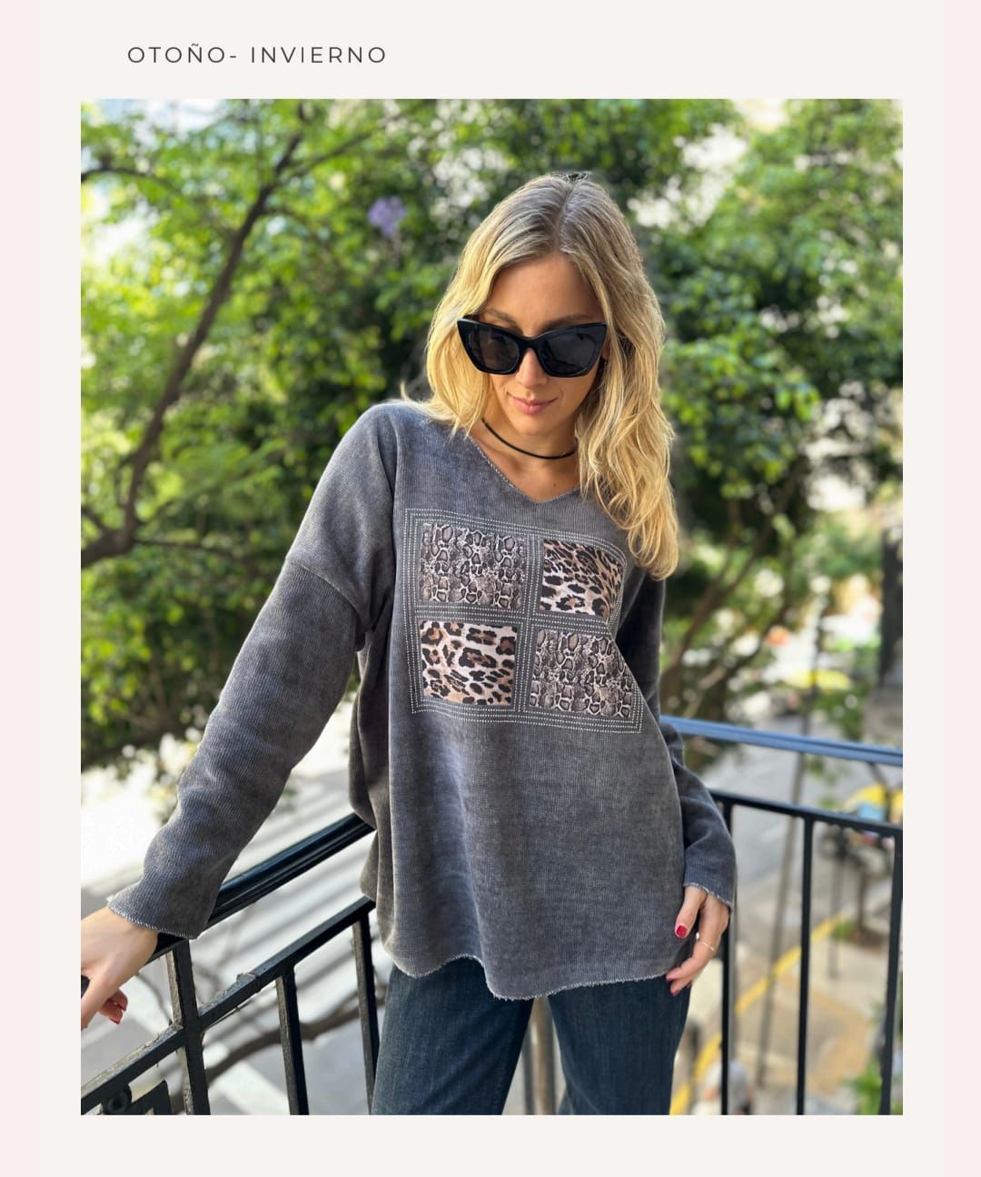 Mujer con gafas de sol y un jersey gris con estampado de leopardo, de pie al aire libre en un balcón.