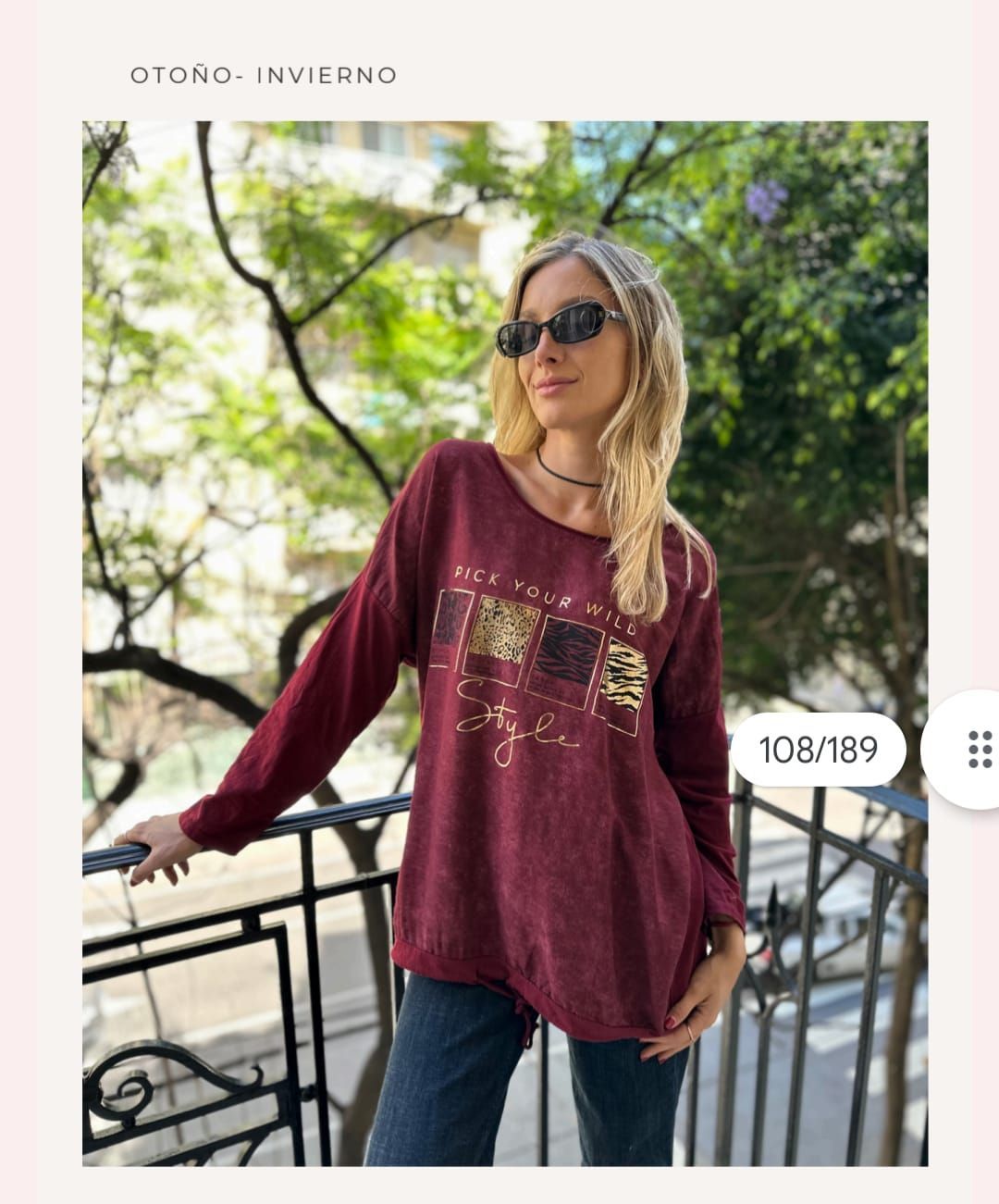 Mujer con gafas de sol, blusa estampada color granate y vaqueros, de pie en un balcón con árboles detrás.