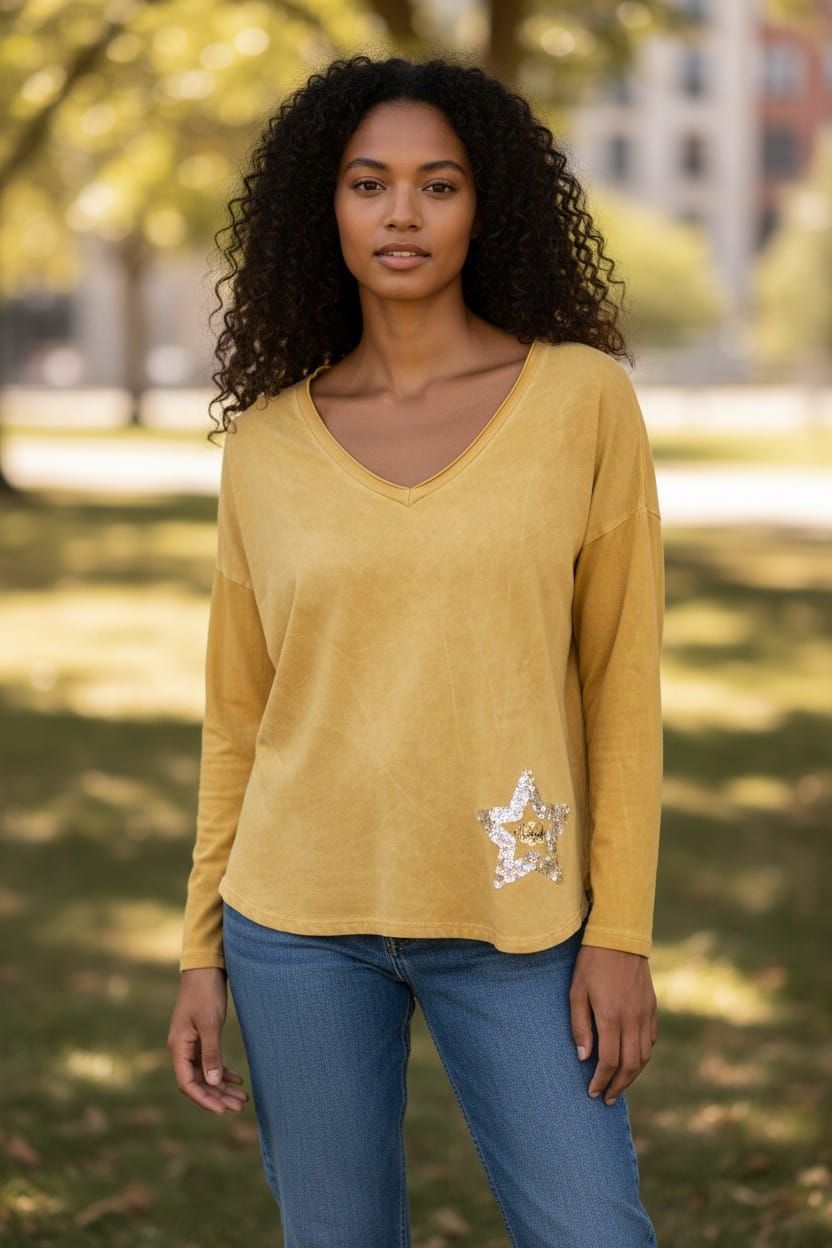 Persona de pie al aire libre con una blusa color mostaza y pantalones vaqueros, con árboles difuminados al fondo.