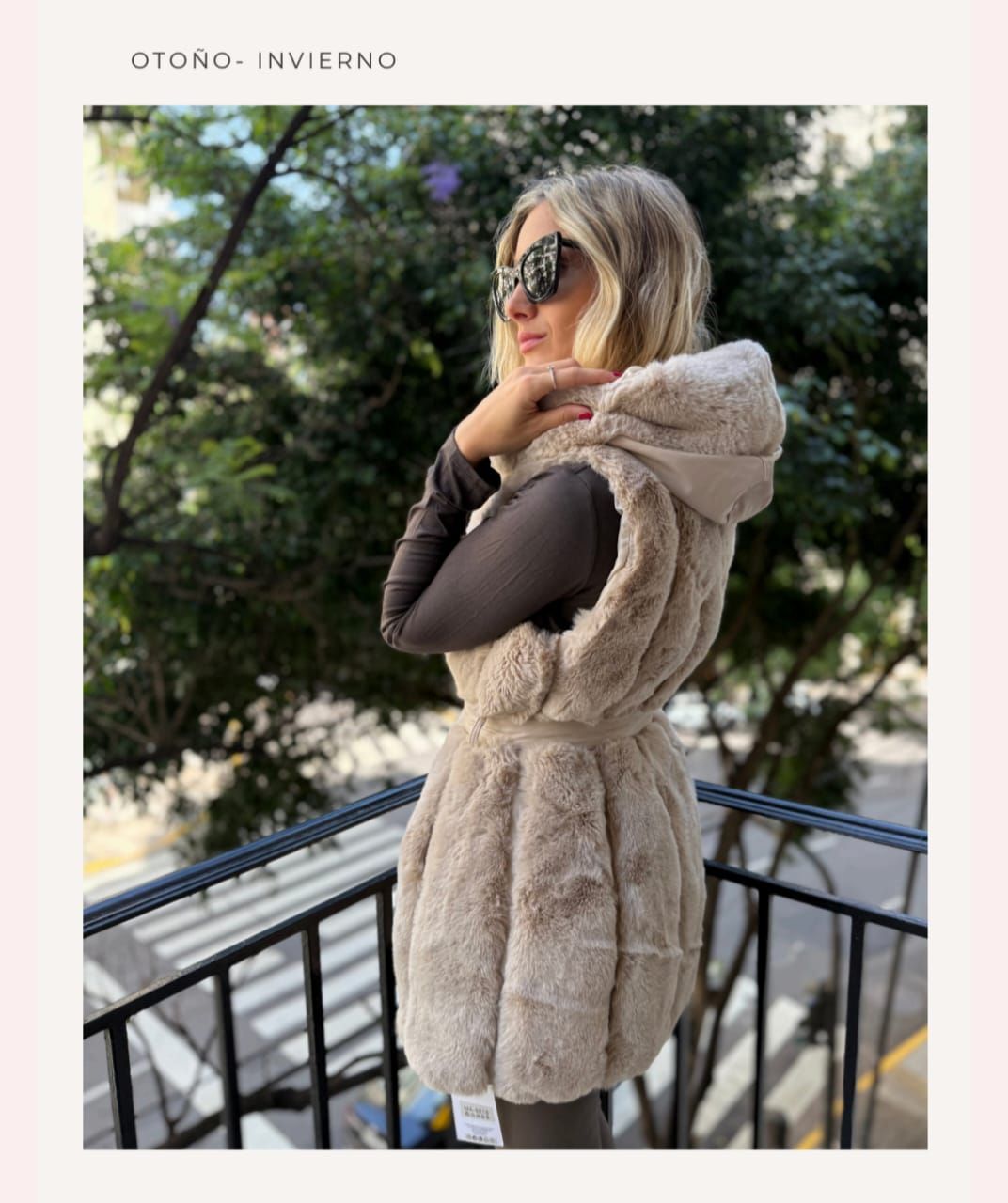 Mujer con abrigo de piel beige y gafas de sol de pie en un balcón en un entorno exterior frondoso.