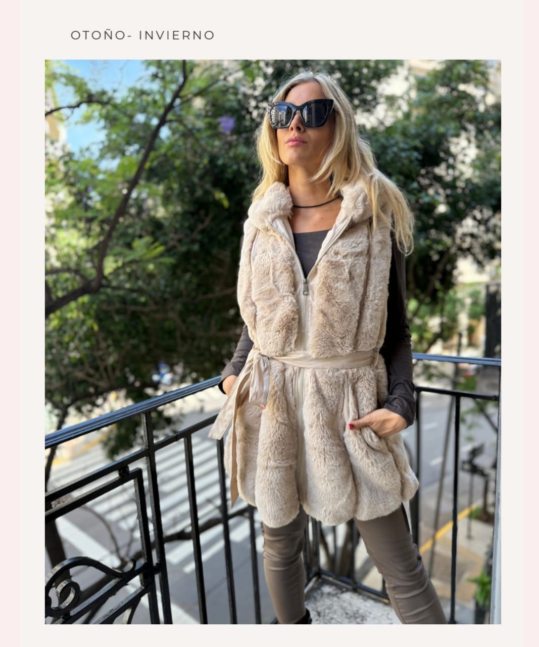 Mujer con abrigo de piel beige y gafas de sol de pie en un balcón en un entorno exterior frondoso.