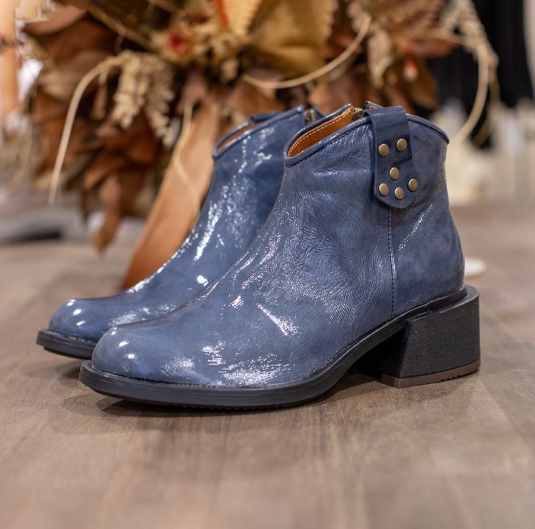 Botines de ante azul con tacones de bloque negros sobre un suelo de madera, con un arreglo floral al fondo.
