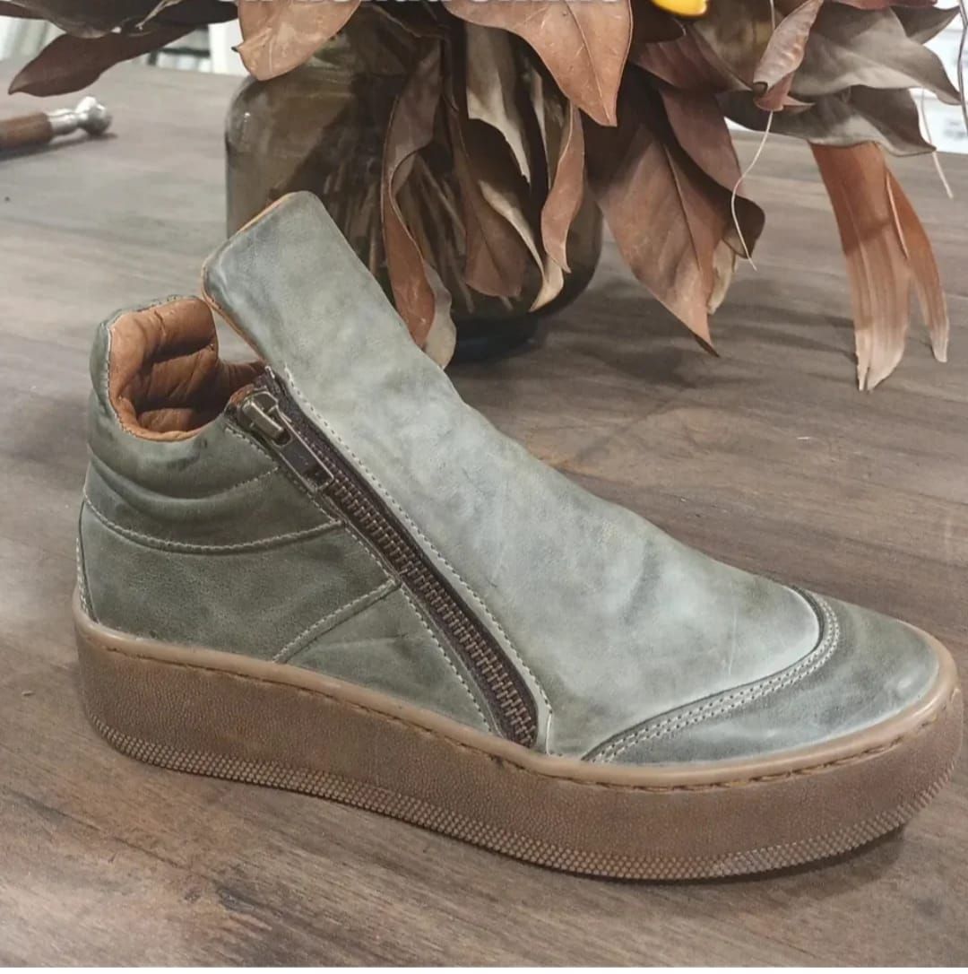 Botín verde sin cordones con cremallera lateral y suela color canela sobre un suelo de madera.
