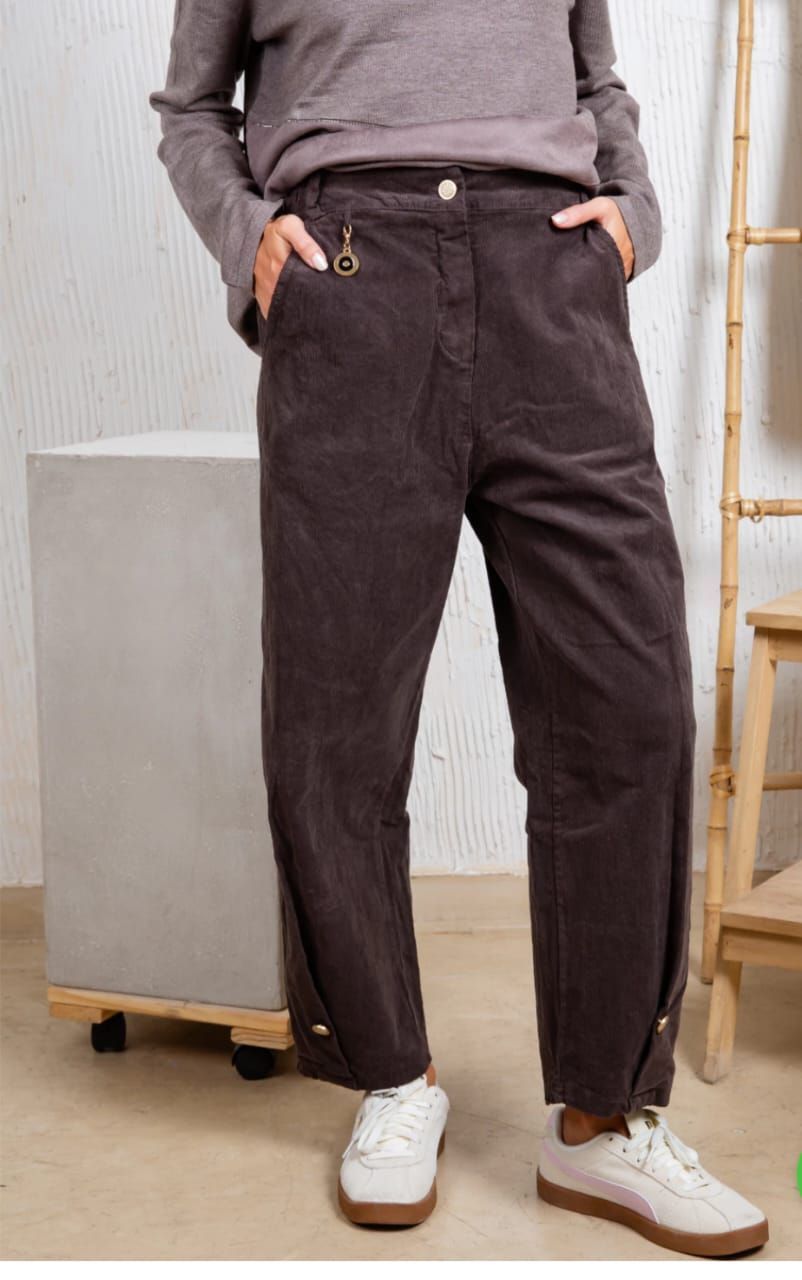 Persona vestida con pantalones marrón oscuro y zapatillas blancas, con las manos en los bolsillos, de pie en un estudio.