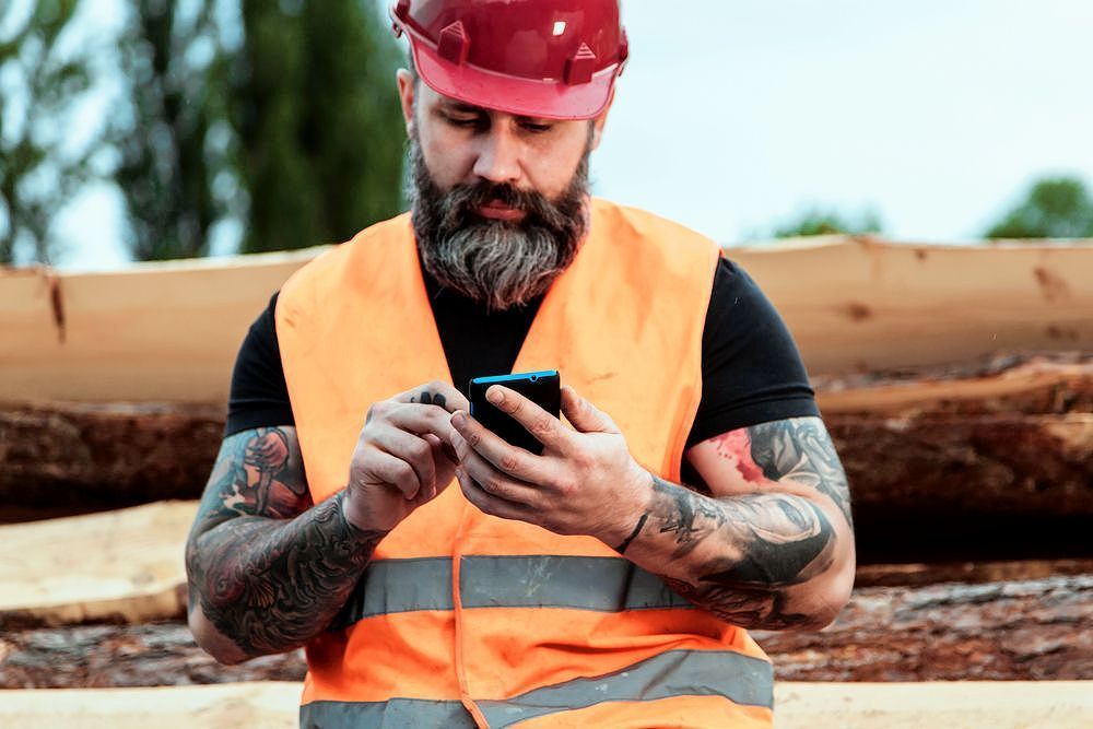 A construction worker is sitting on a pile of logs looking at his cell phone. A construction worker is sitting on a pile of logs looking at his cell phone.