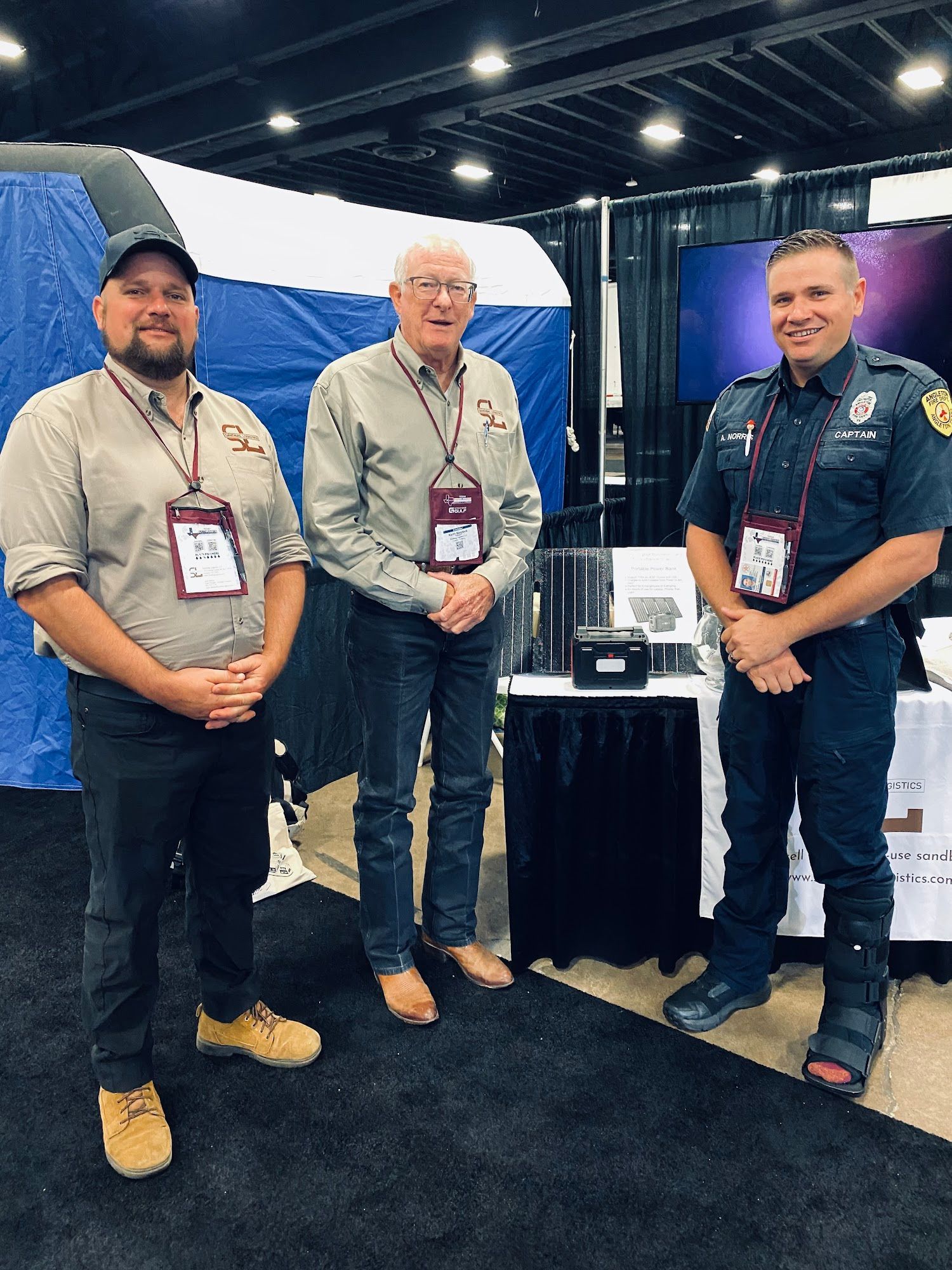 Three men standing at a trade show booth beside a table with equipment and blue backdrop