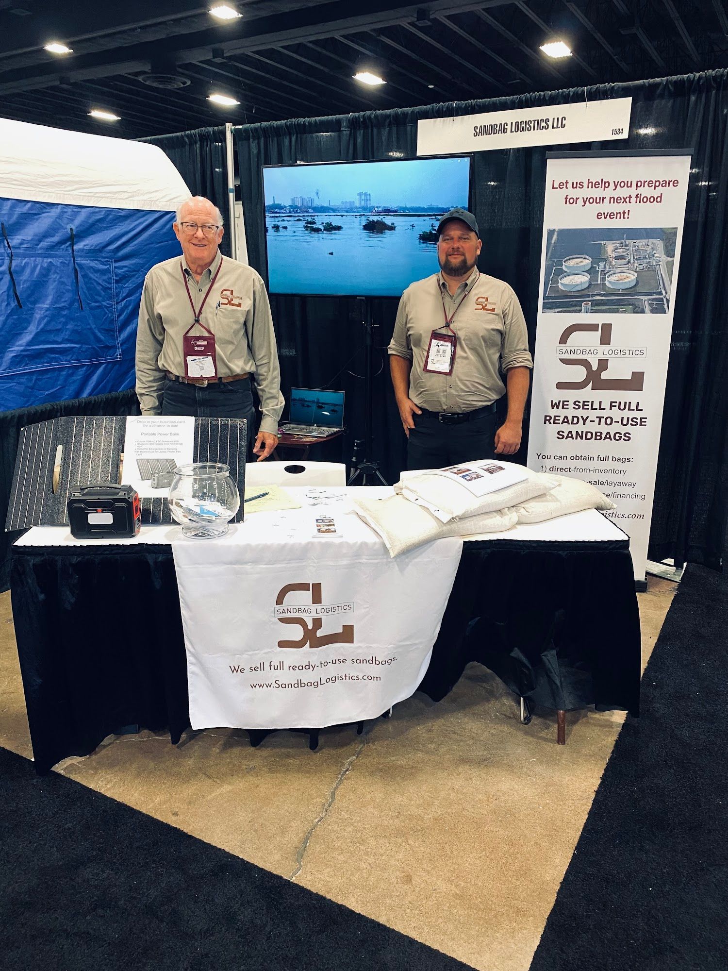 Two men behind a trade show booth with banners, table displays, and a TV screen showing a presentation