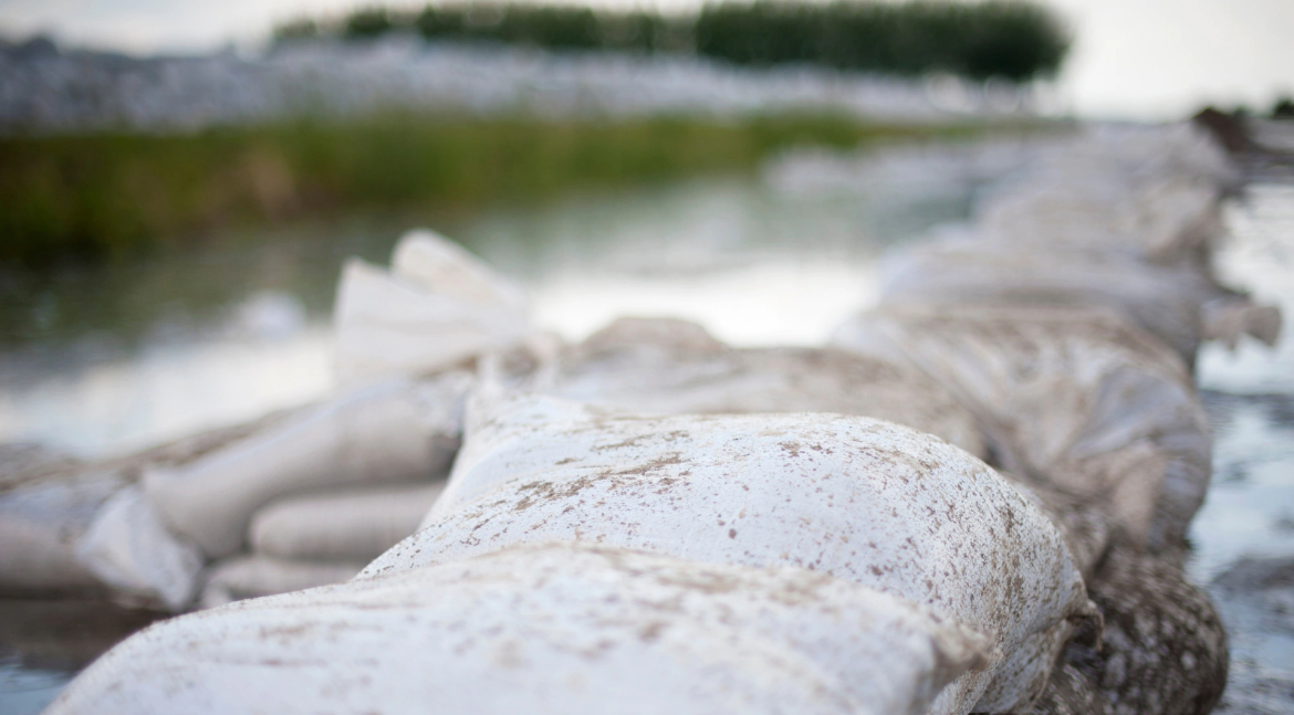 Large white sandbags lining a riverbank beside calm water and greenery