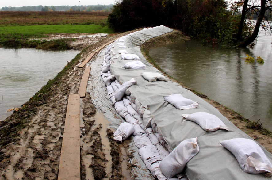 Sandbagged flood barrier curving beside flooded fields and a waterway under overcast skies