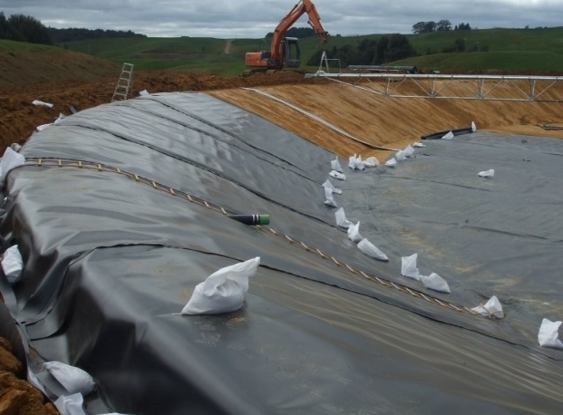 Black liner in excavated pond, with sandbags and excavator on the far bank under cloudy sky