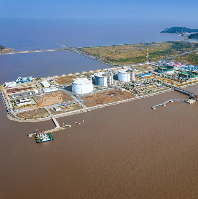 Aerial view of an industrial waterfront with storage tanks, docks, and brown water beside a coastal shoreline