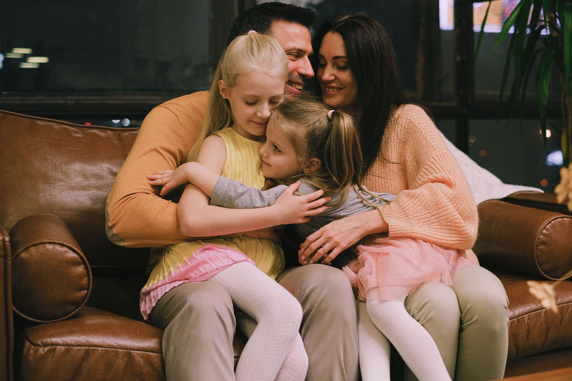 Family sitting close together on a brown couch, smiling and hugging in a cozy living room.