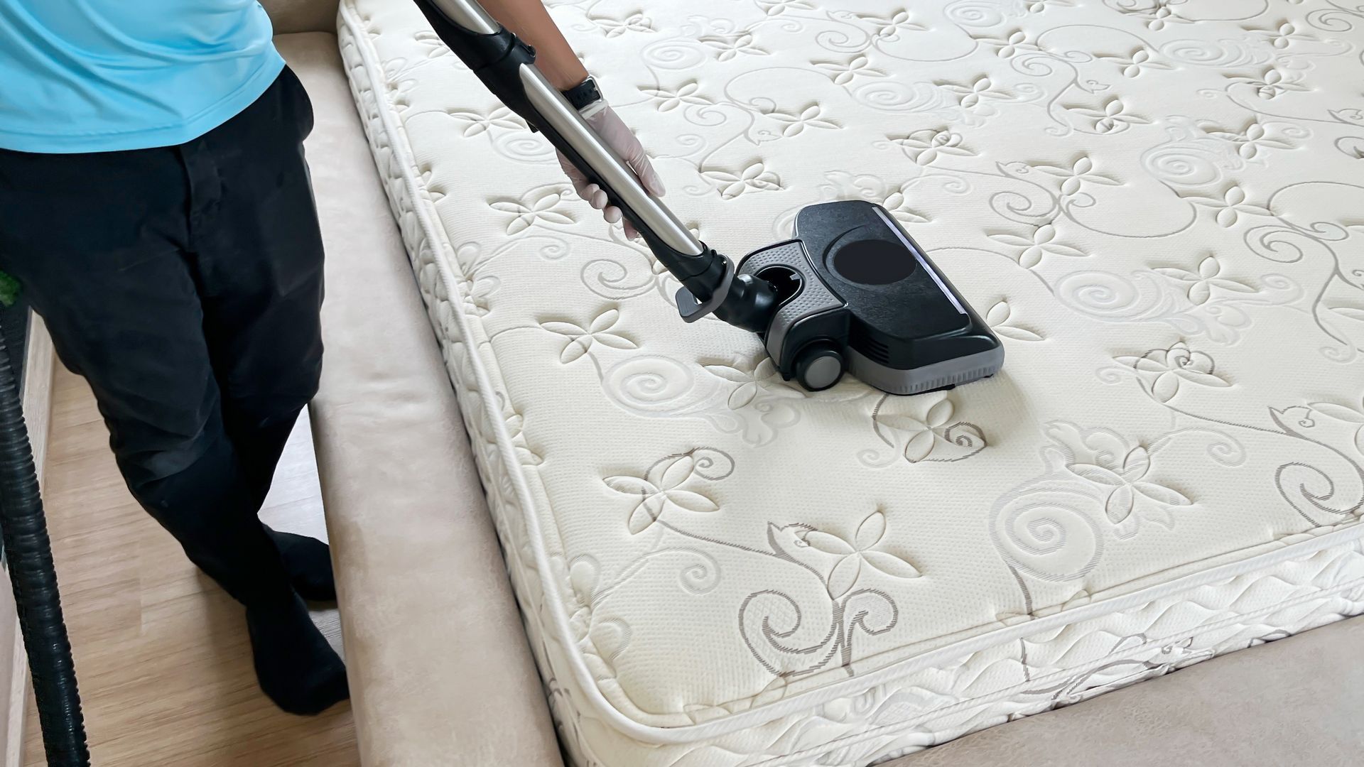 Person vacuuming a cream-colored mattress. The person is wearing gloves, a light blue shirt, and black pants.