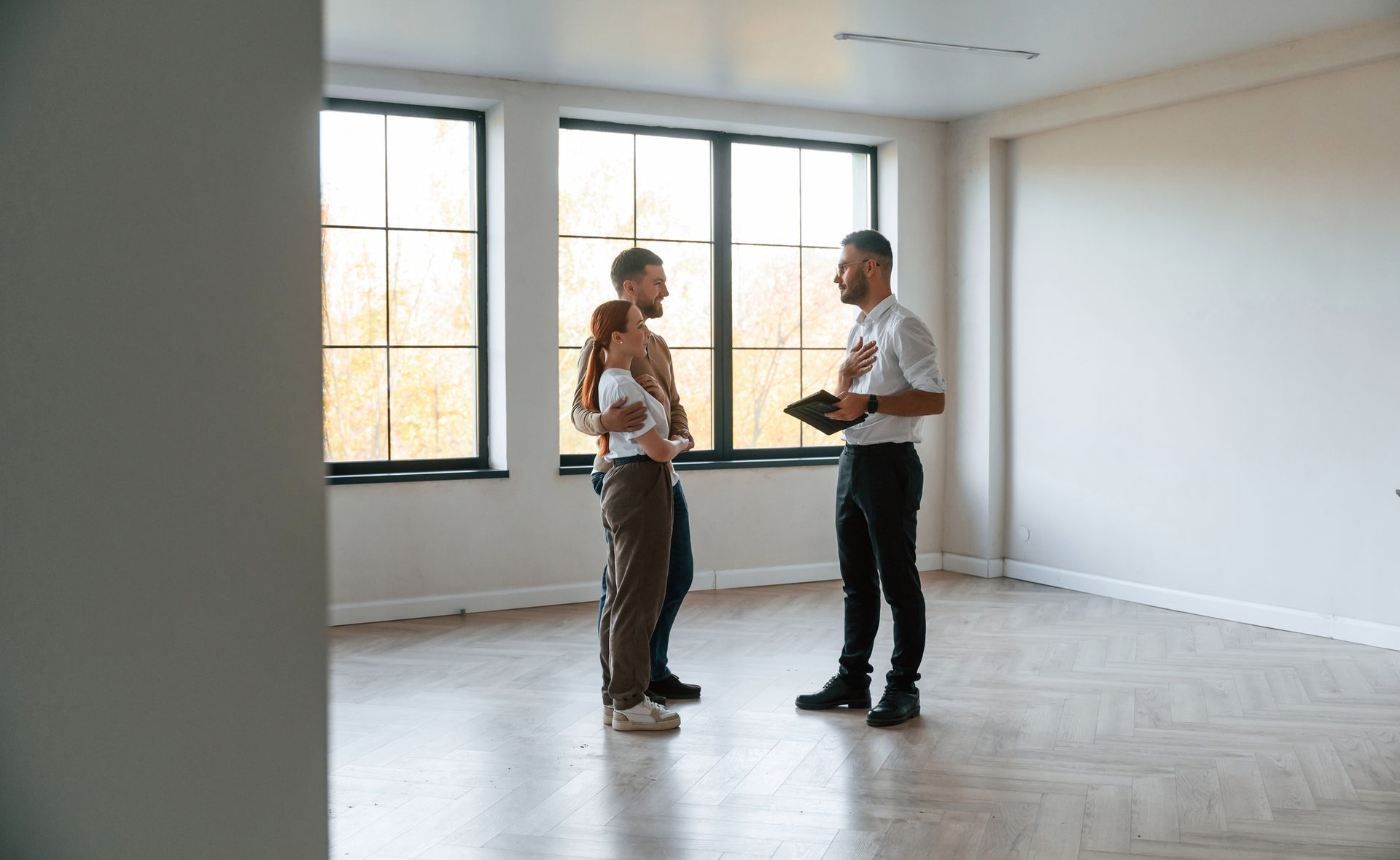 A man and a woman are standing in an empty room talking to a real estate agent.