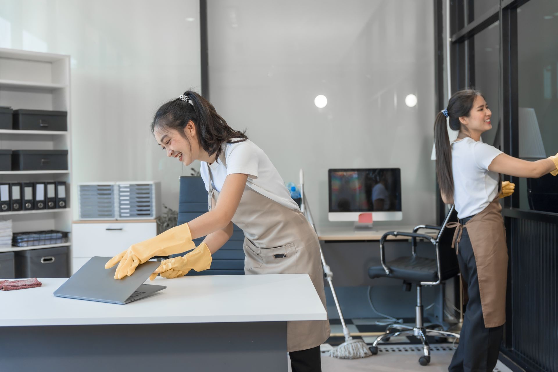 Two women cleaning an office; one wipes a window, the other cleans a laptop.