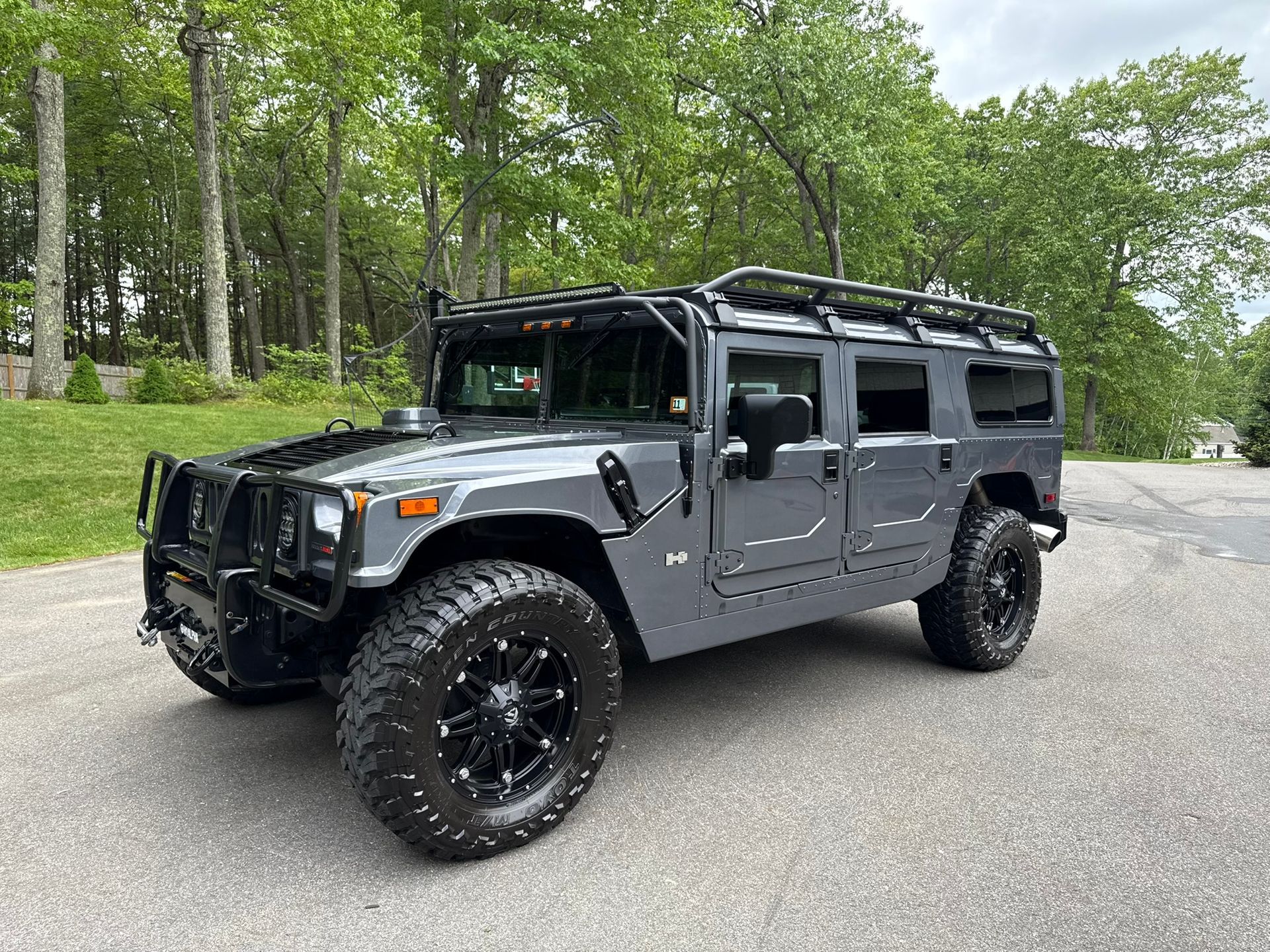 Gray Hummer SUV parked on pavement with black wheels and roof rack, trees in background.