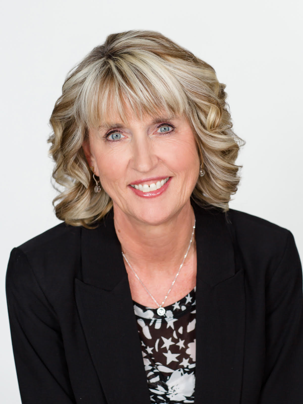Woman smiling, wearing a black blazer over a patterned blouse. Blond hair, neutral background.