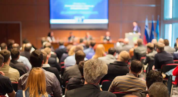 A large group of people are sitting in a conference room watching a presentation.