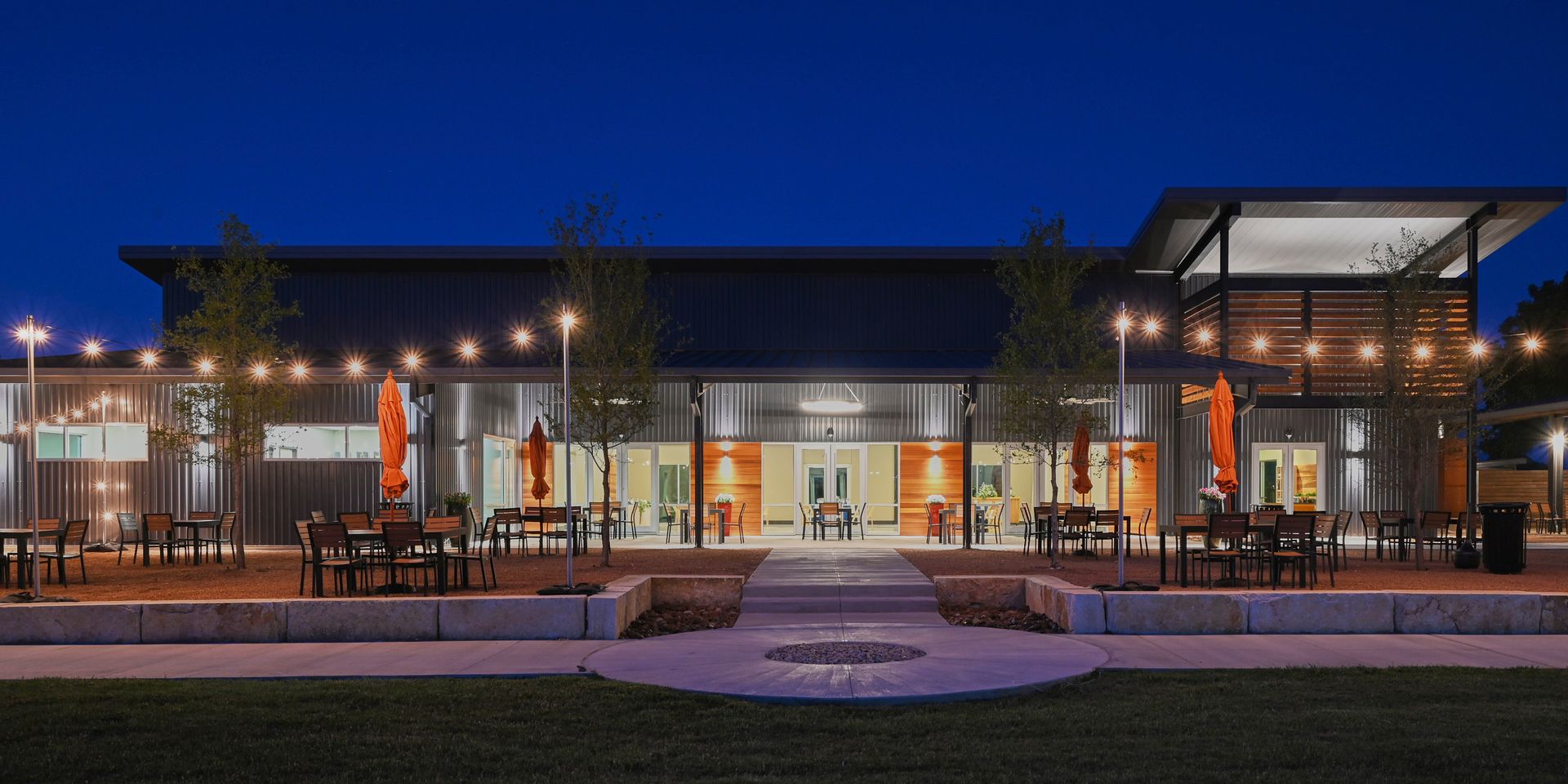 Modern building with lit glass facade at dusk and outdoor seating in front