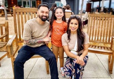 Three people sit on wooden chairs outdoors, smiling at the camera.