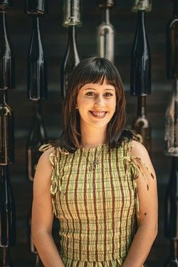 Smiling woman in a sleeveless plaid dress standing before hanging black bottles and glassware