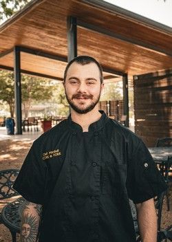 Chef in a black uniform standing outdoors under a wooden patio, smiling at the camera.