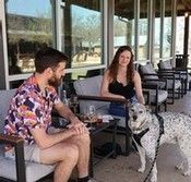 Two people sitting at an outdoor café with a black-and-white dog standing nearby