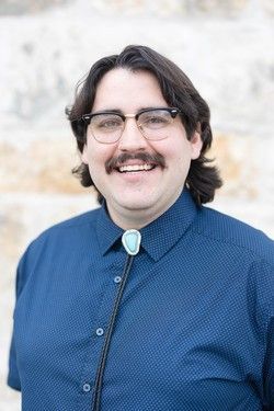 Smiling person with glasses and mustache wearing a blue shirt and bolo tie against a light background