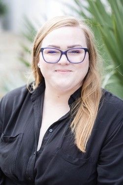 Person with long blonde hair and blue glasses, wearing a black shirt, outdoors with blurred greenery background