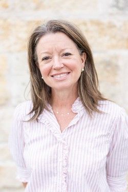 Smiling woman in a light pink blouse standing against a softly blurred stone wall.