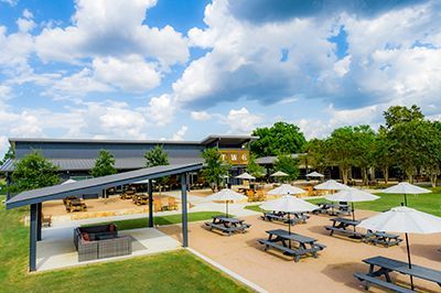 Outdoor patio with picnic tables and umbrellas beside a covered seating area under a partly cloudy sky