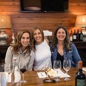 Three smiling friends seated at a wooden restaurant table with wine glasses and plates