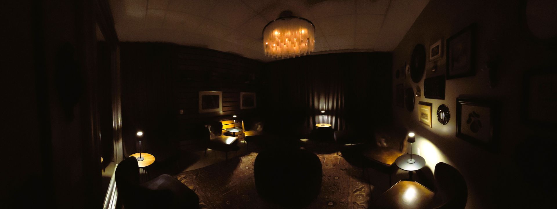 Dimly lit room with a round table, warm lamps, framed wall art, and a glowing pendant light above.