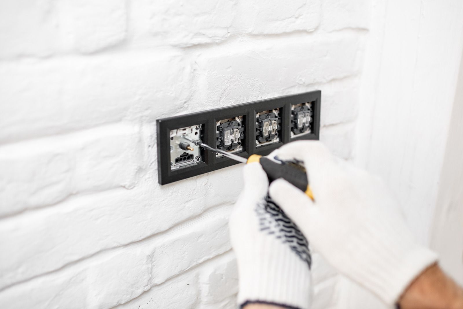 Gloved hands installing a black electrical outlet plate on a white brick wall