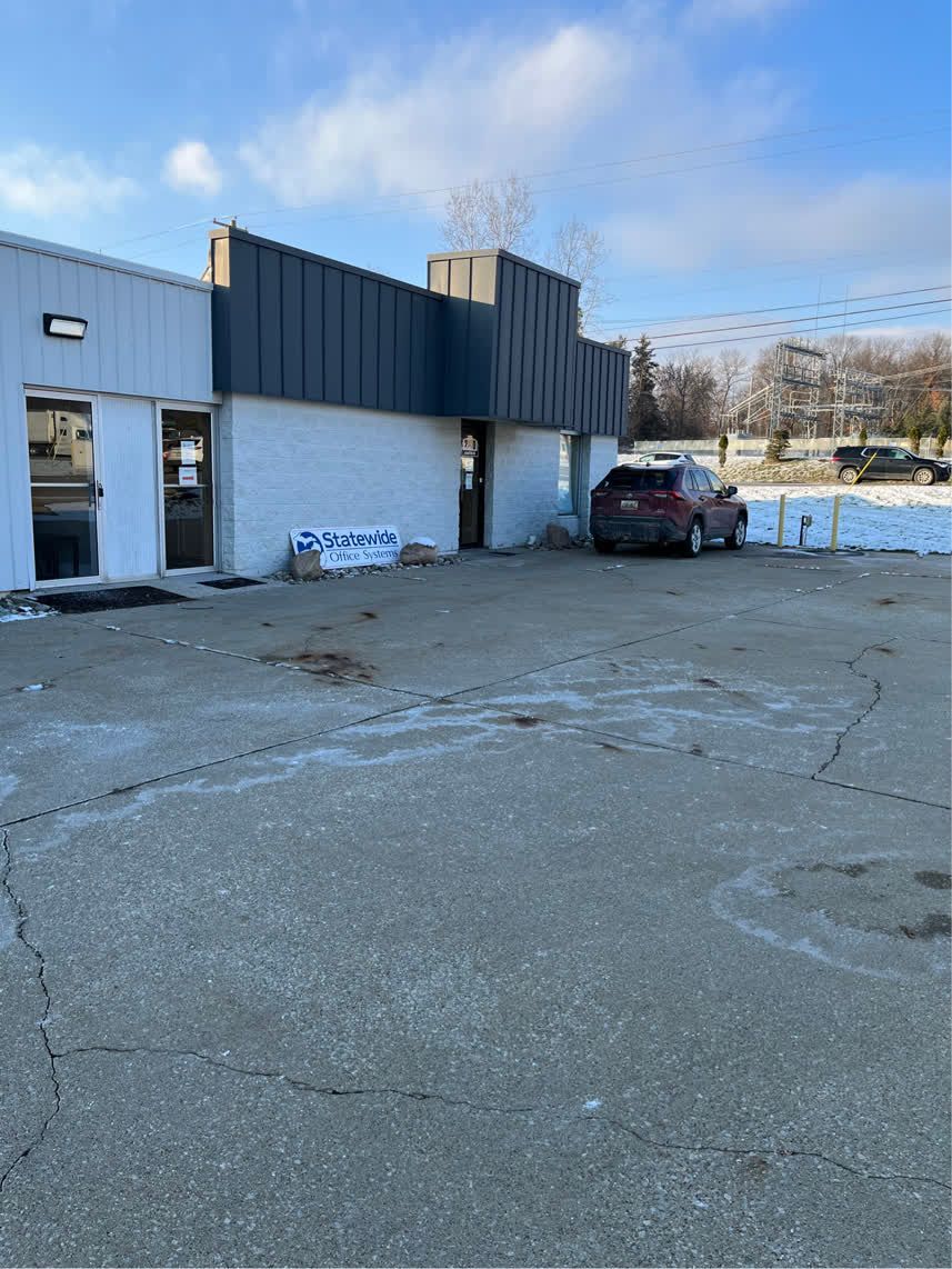 Exterior of a white commercial building with a dark gray roof. A red car is parked on the gray asphalt.