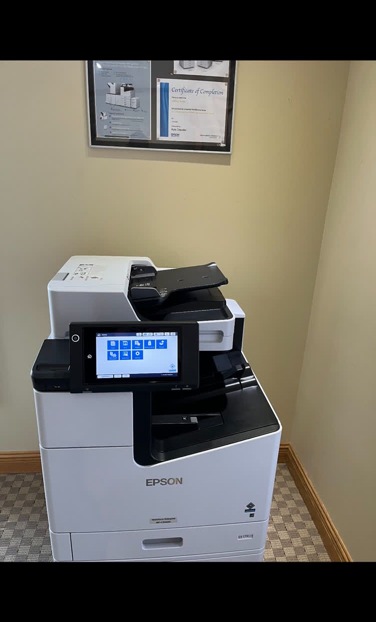 Multifunction printer with a blue-lit screen, sitting under framed documents against a beige wall.